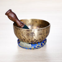Full Moon Jhumkana singing bowl with wooden mallet and blue brocade cushion, showing fire-spot patina and laser-carved symbol — handcrafted in Nepal